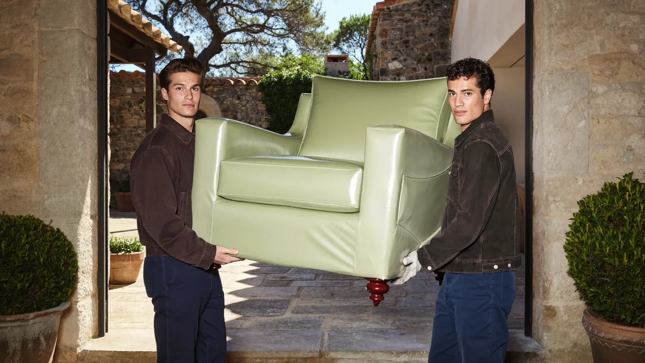 Deux déménageurs portant avec soin un fauteuil emballé dans une maison de village en pierre